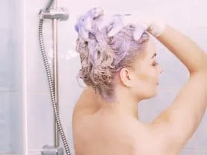 Woman washing toned blonde hair with purple shampoo in the shower to reduce brassiness and maintain cool blonde color